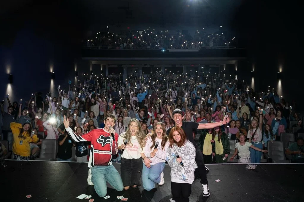 Harper Zilmer on stage with four other people, all smiling and posing for a group photo with a large, enthusiastic audience behind them in a dark theater. Audience members are holding up their phone flashlights, adding a bright, celebratory atmosphere to the scene.